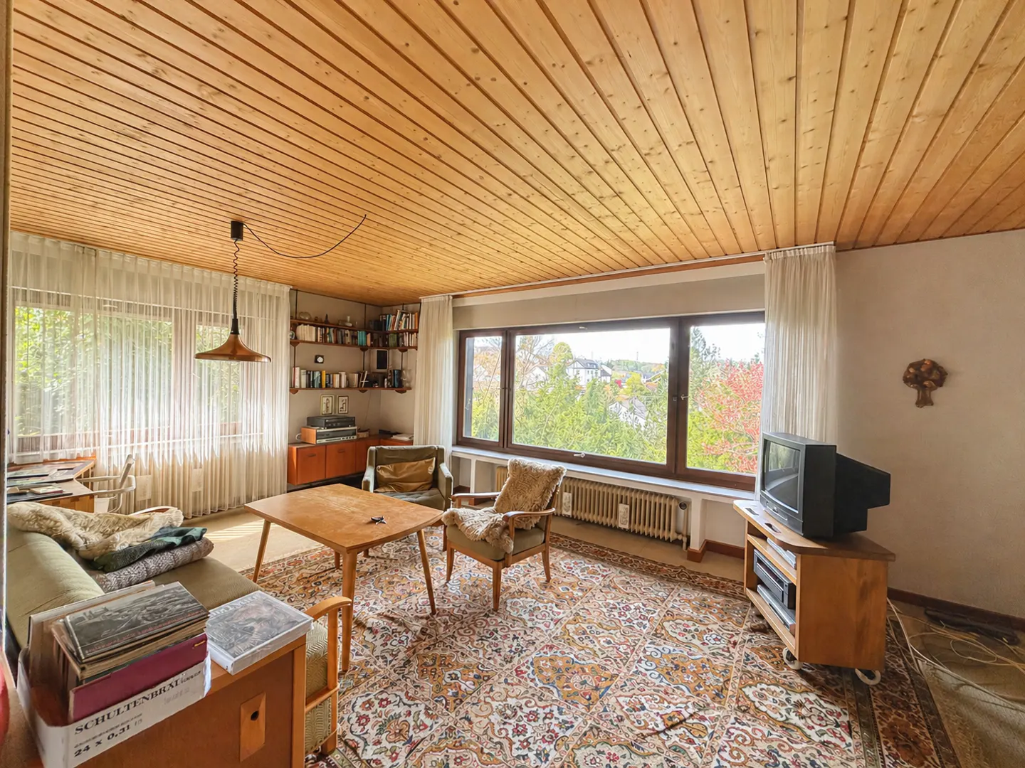 Living room with wood-paneled ceiling, large windows, patterned rug, sofa, chairs, and vintage TV. Bookshelves line one wall.