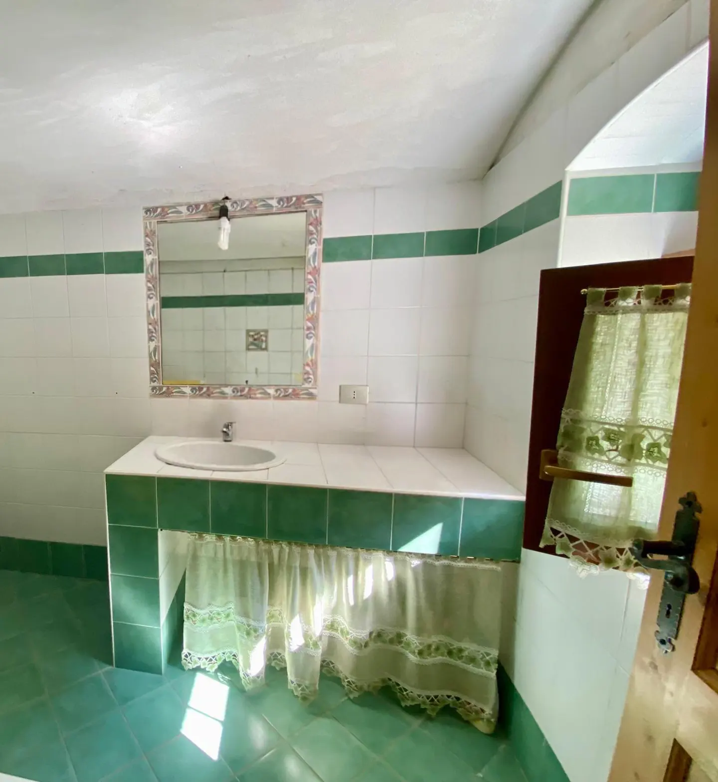 Bathroom with green tile floor and trim, white walls, sink, mirror, and window with sheer curtain.