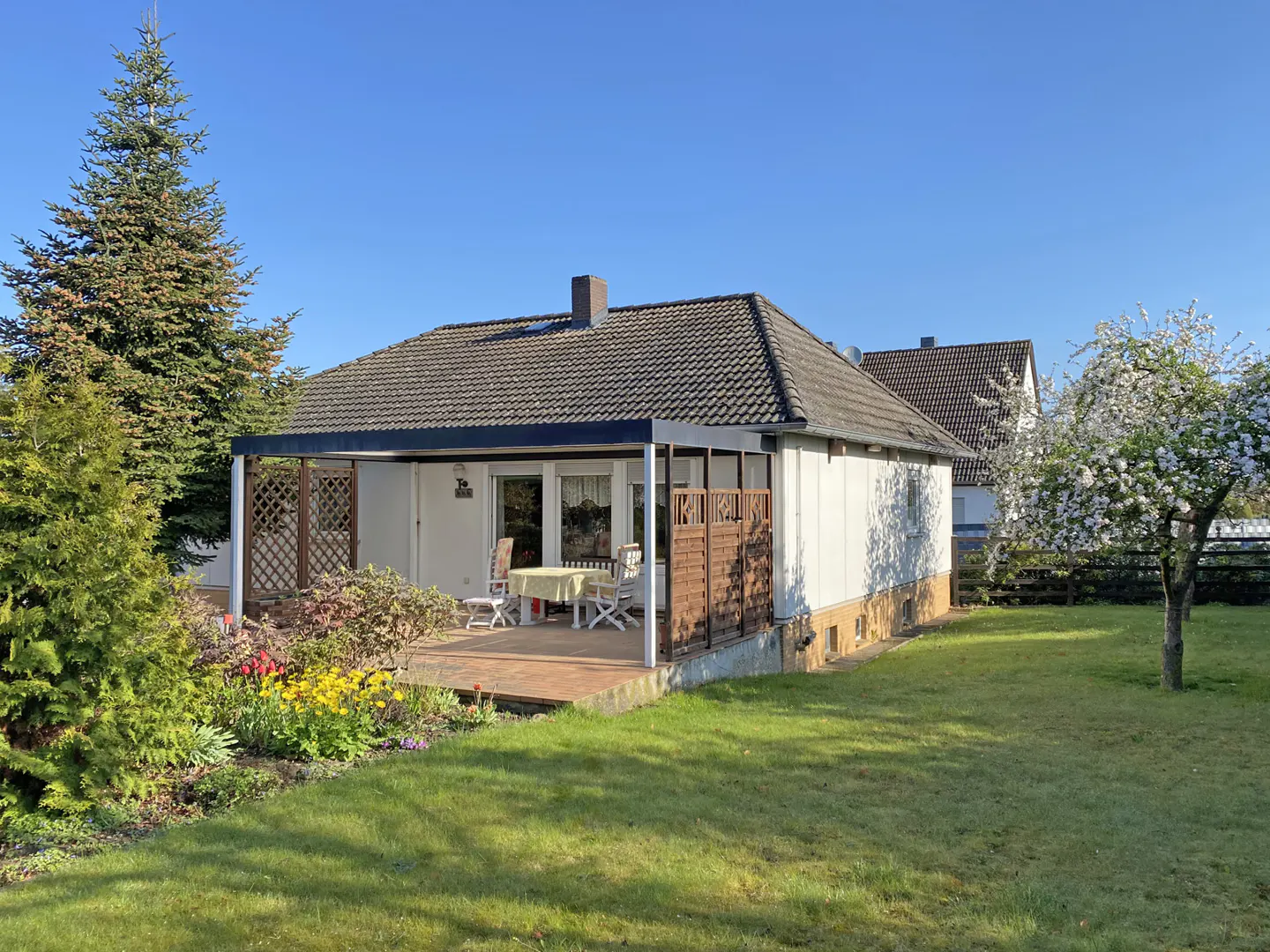 A white house with a brown roof and a patio with a table and chairs sits on a green lawn.