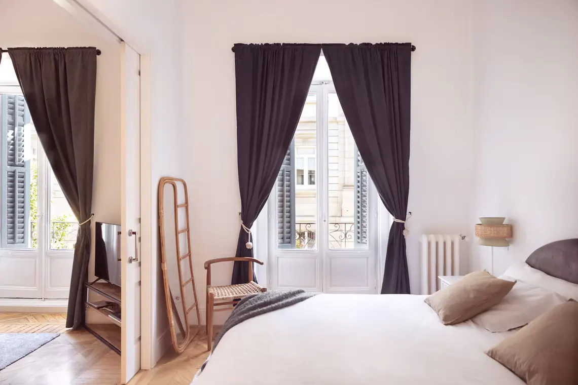 Bright bedroom with white walls, a bed with pillows, and dark curtains framing a window with blue shutters. A wooden chair and mirror are nearby.