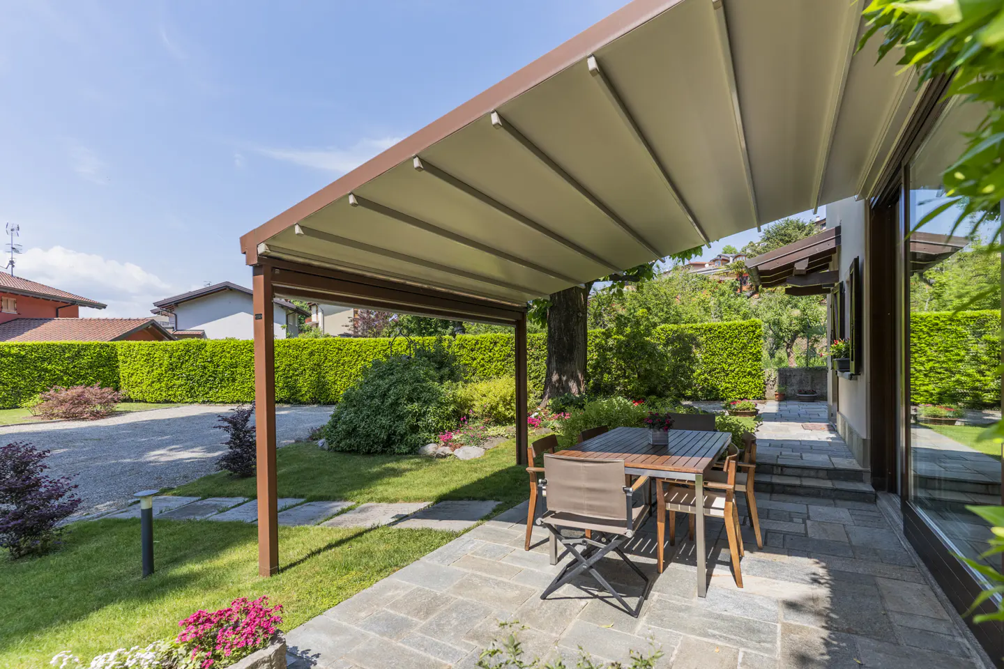 Outdoor patio with a brown pergola, a wooden table and chairs on a stone patio, and a green lawn.