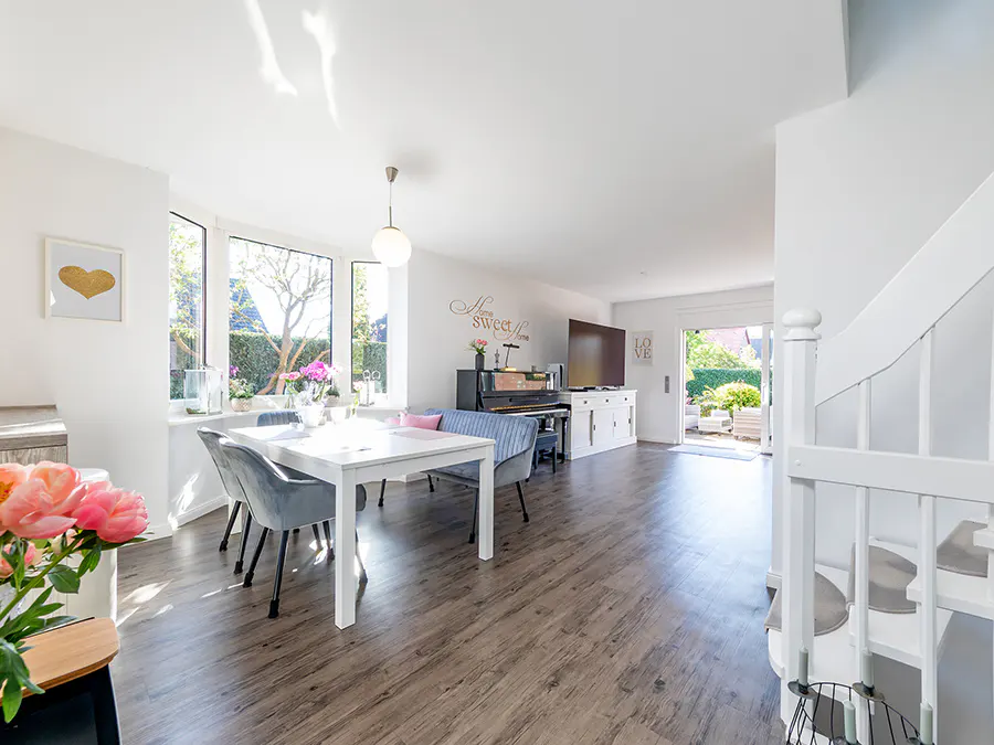 Bright, open-concept living space with wood floors, white walls, dining table, piano, TV, and stairs. Natural light floods the room.