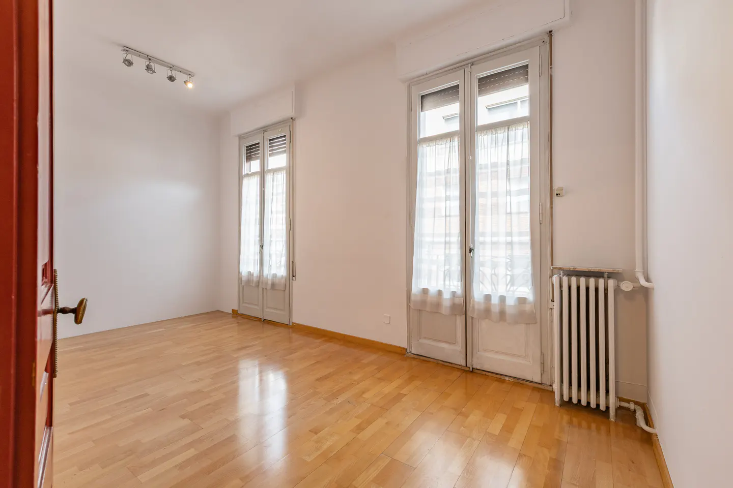 Empty room with light wood floors, white walls, two sets of French doors with sheer curtains, and a white radiator.