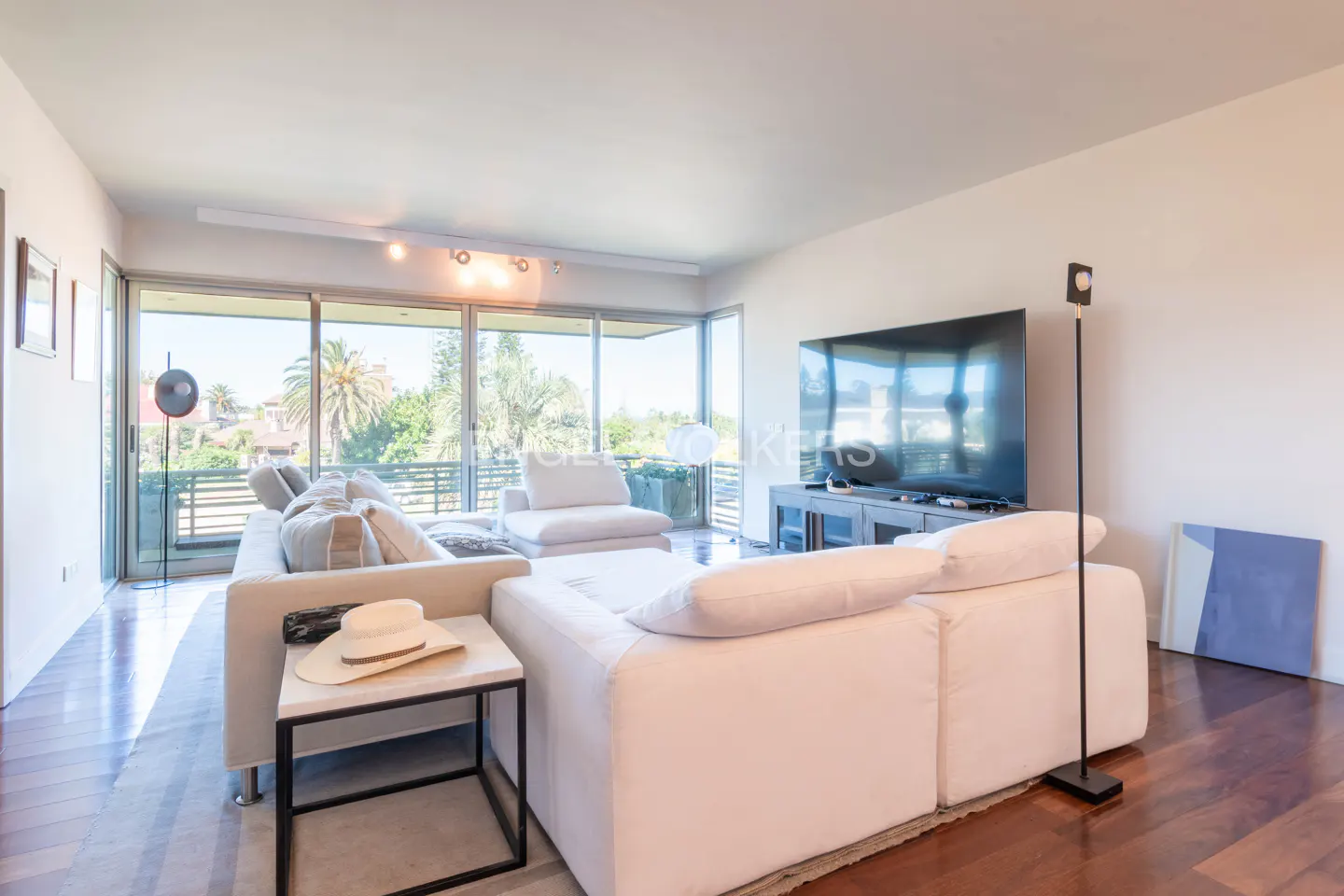 Bright living room with white sectional sofa, marble side table with hat, and large windows overlooking greenery.