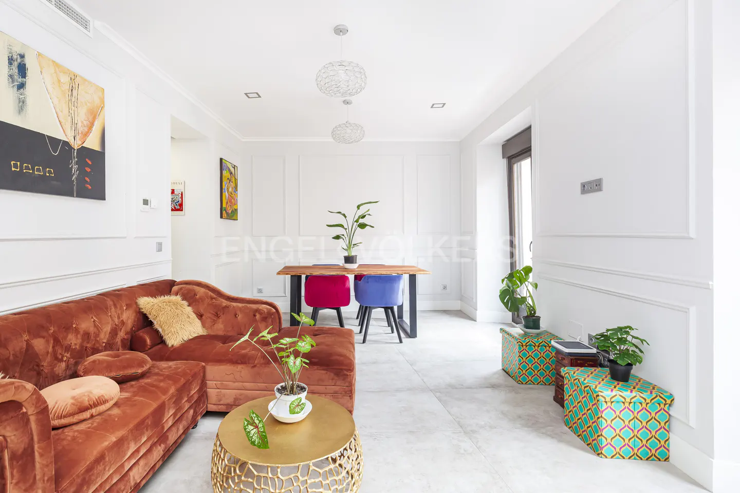 Living room with a brown velvet sofa, gold table, and a wood table with pink and blue chairs. White walls and light gray floors.