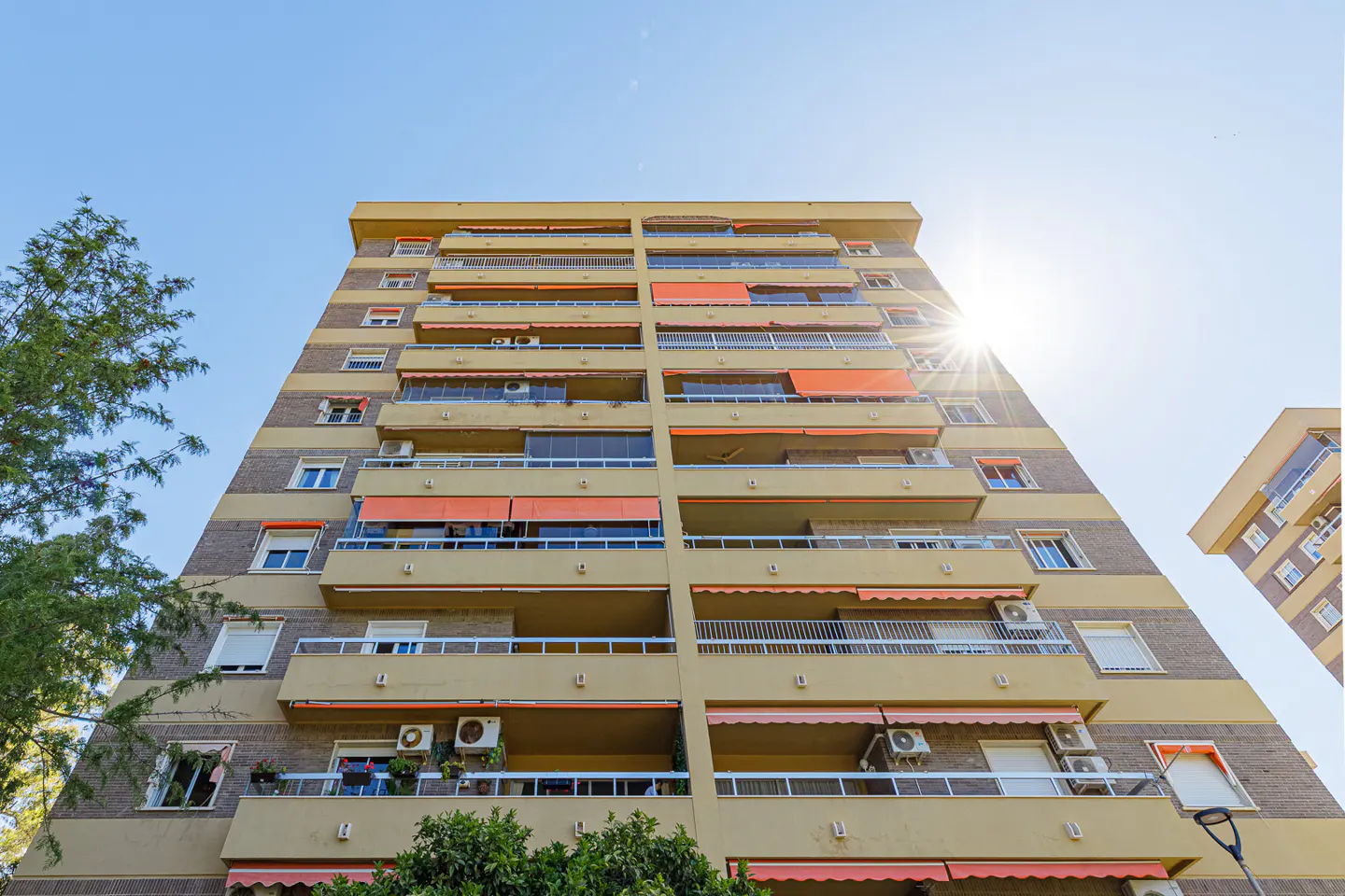 Exterior view of a tall apartment building with balconies and orange awnings under a bright blue sky.