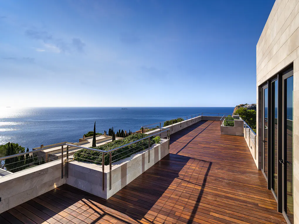 Wide, sunny, wood-planked deck with ocean view. Railings, planters, and a stone building with black-framed windows are visible.