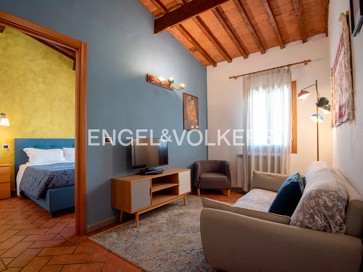 Cozy living room with blue accent wall, terracotta floors, and exposed wood ceiling. A bedroom is visible through an open doorway.