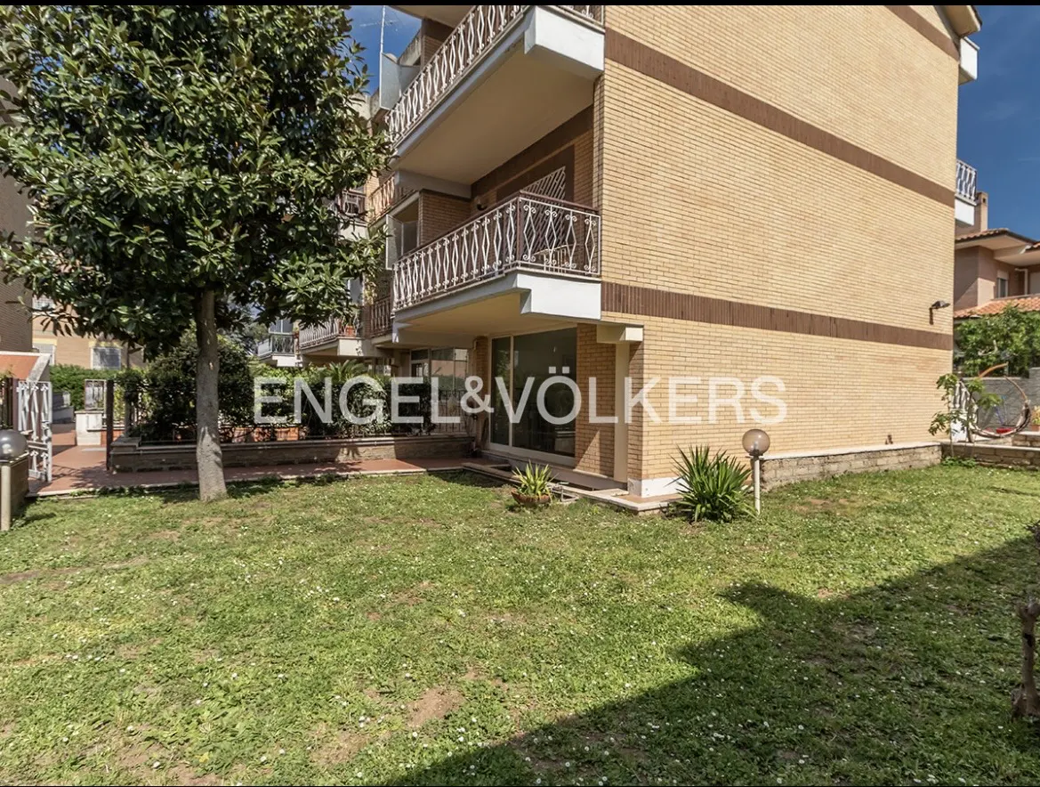 Exterior view of a tan brick apartment building with balconies and a green lawn. The Engel & Völkers logo is visible on the building.