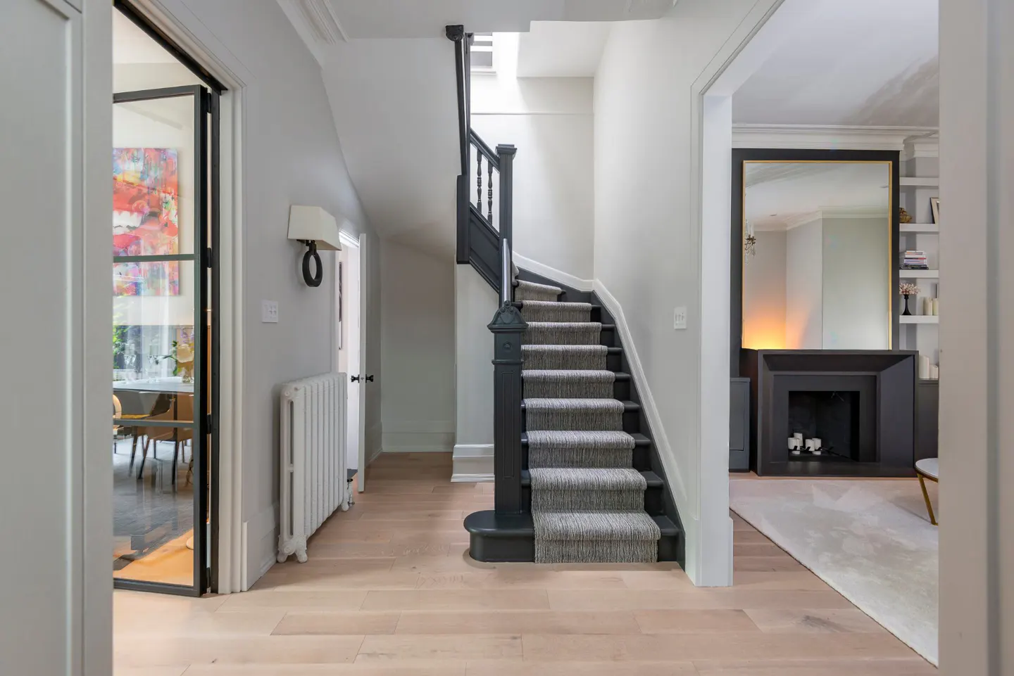 Hallway with light wood floors, gray walls, and a black staircase with gray carpet. A dining room and living room are visible through doorways.