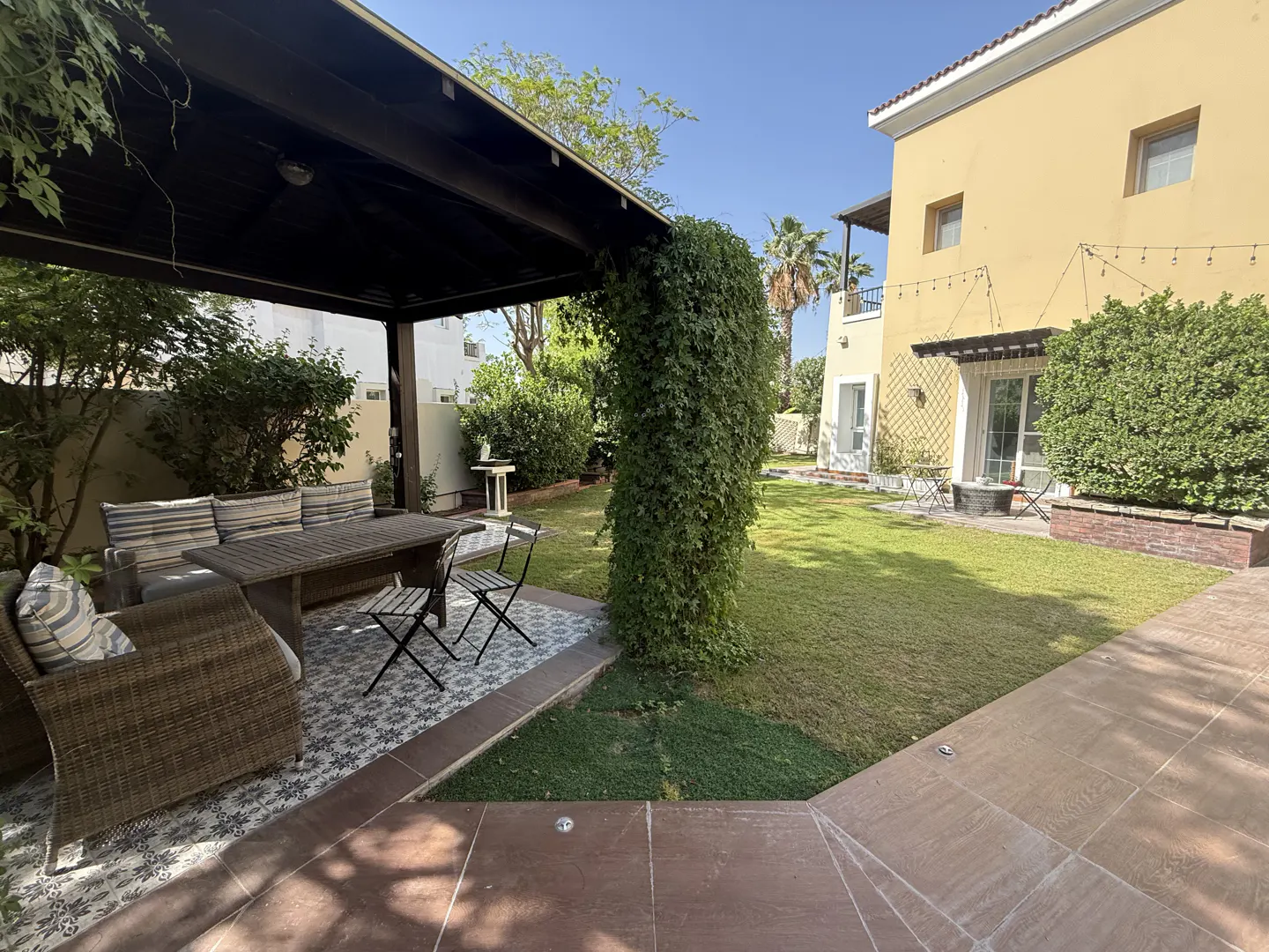 A backyard with a covered patio, a table and chairs, and a green lawn. A yellow house is in the background.
