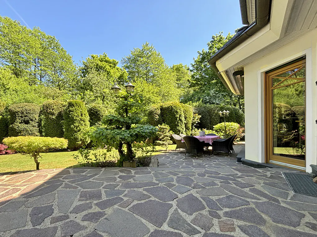 Stone patio with outdoor table and chairs, surrounded by green trees and bushes, next to a white house with a wood-framed door.