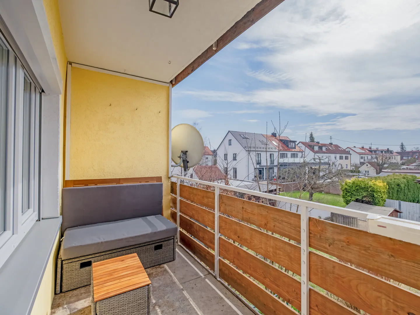Balcony with gray cushioned bench, small table, and wood railing overlooking a residential area under a cloudy sky.