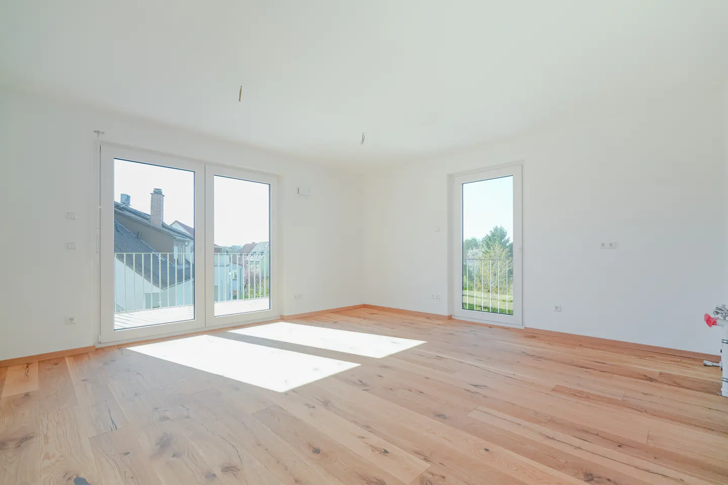 Bright, empty room with light wood floors and white walls. Two sets of glass doors lead to balconies with views of trees and houses.