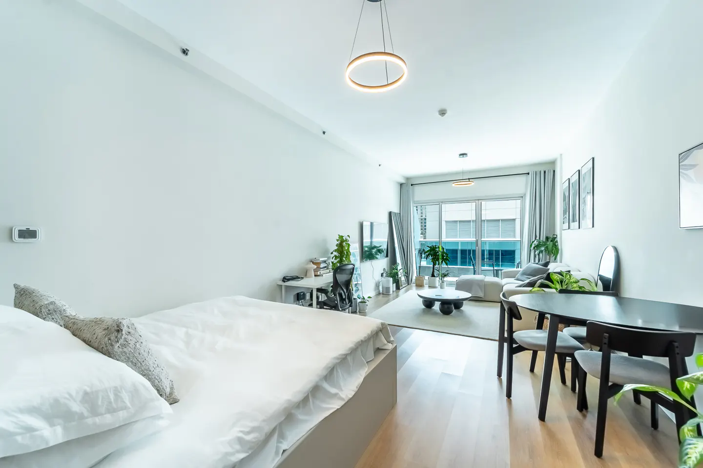 Bright, modern studio apartment with a bed, desk, living area, dining table, and large windows. The color scheme is primarily white and gray.