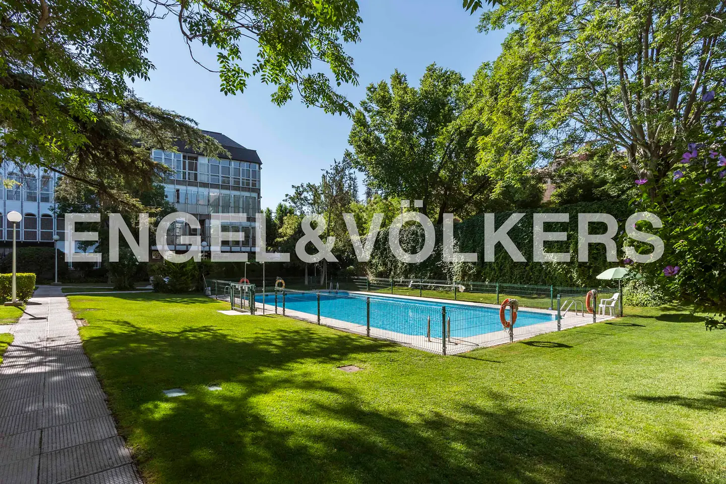 Outdoor swimming pool surrounded by a green lawn, trees, and a modern apartment building under a clear blue sky.