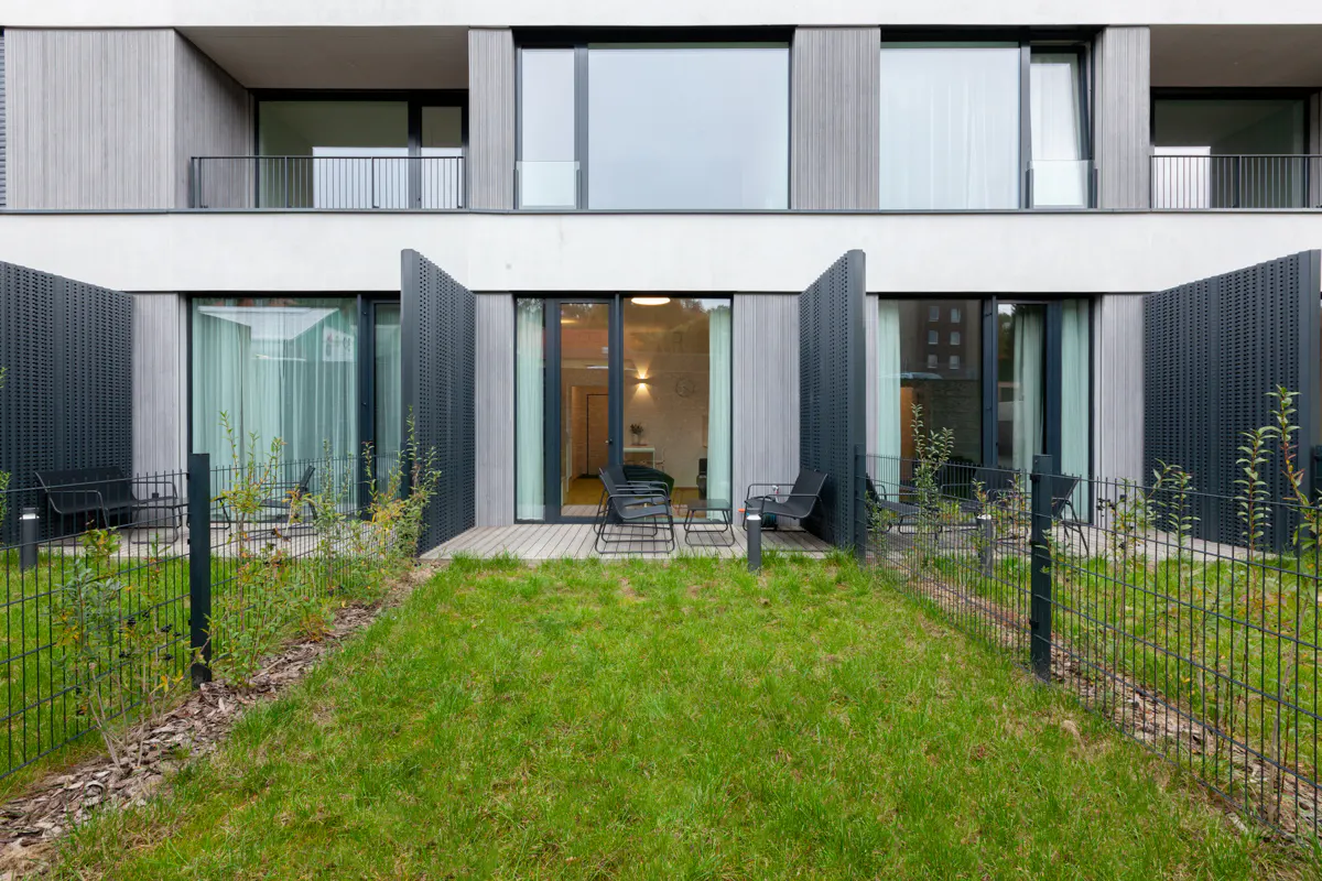 Modern apartment building with private patios. Green lawn, black metal fence, and sliding glass doors. Chairs and tables on wooden patios.