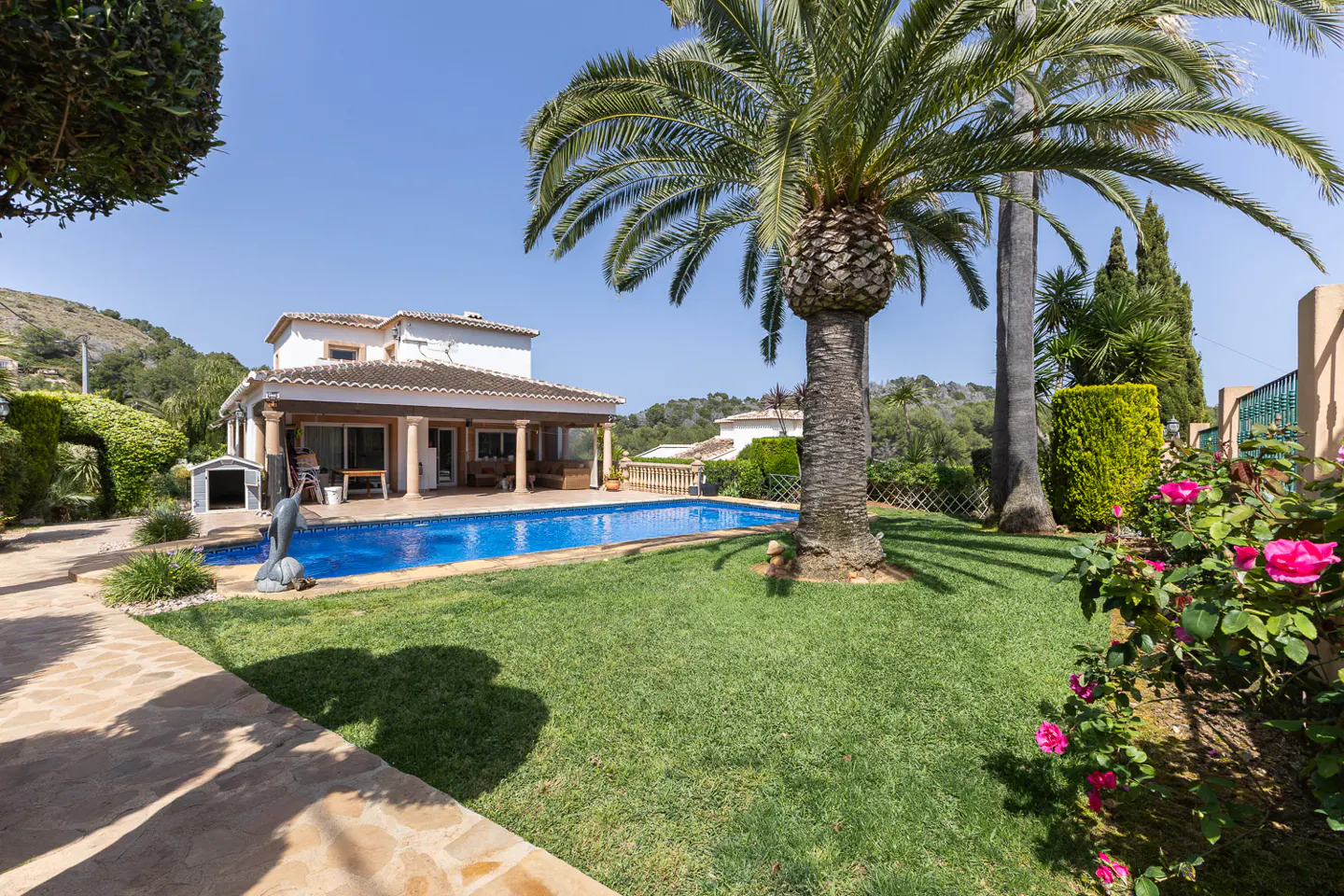 A sunny backyard with a pool, palm trees, and a two-story house with a covered patio. Pink roses are in the foreground.