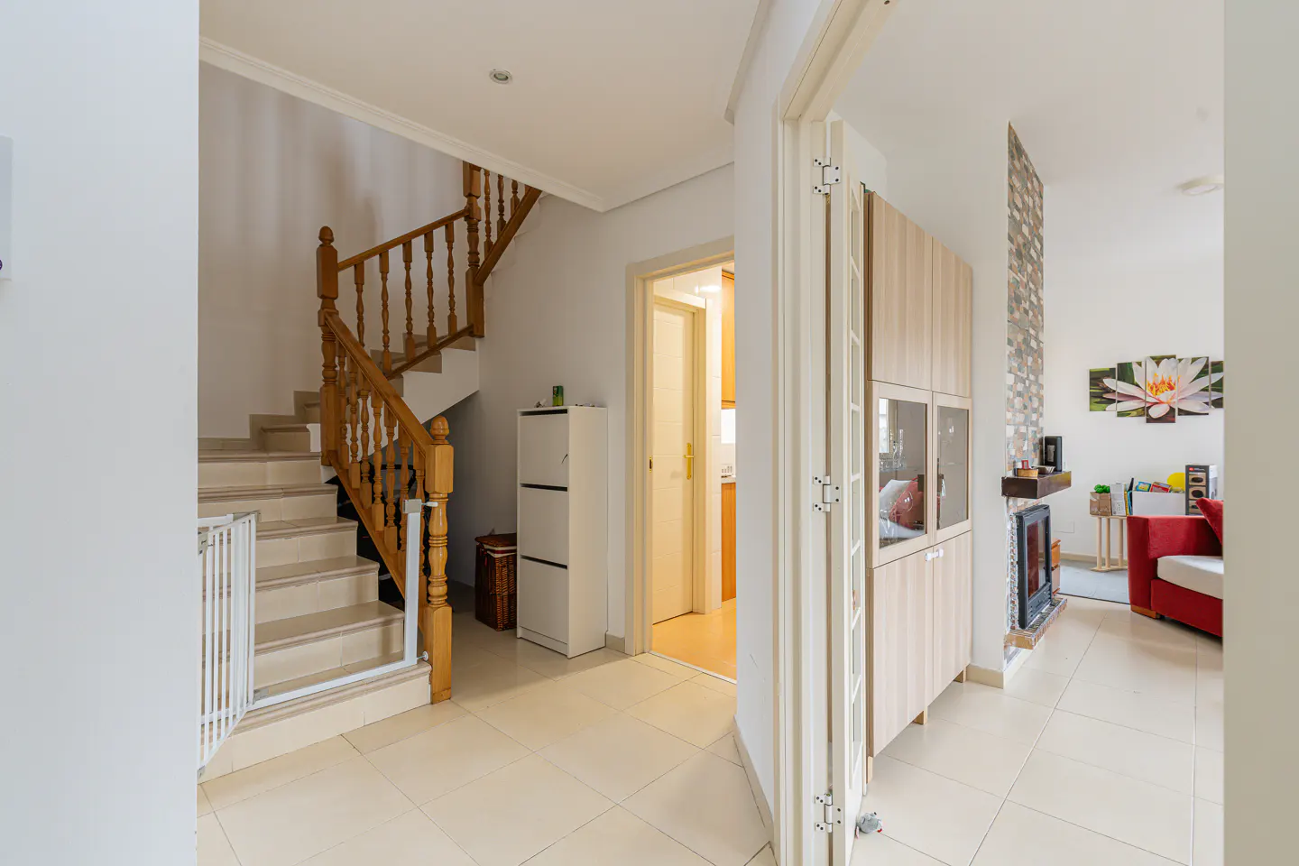 A bright foyer with beige tile floors, a wooden staircase with a white safety gate, and a glimpse into the living room with a red sofa.