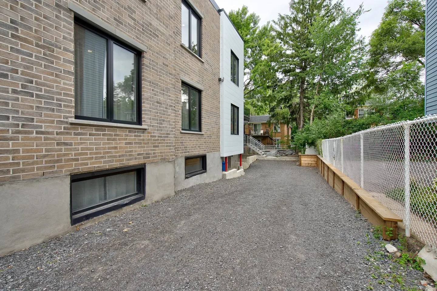 Exterior view of a brick building with black framed windows and a gravel driveway. A chain link fence runs along the side of the property.