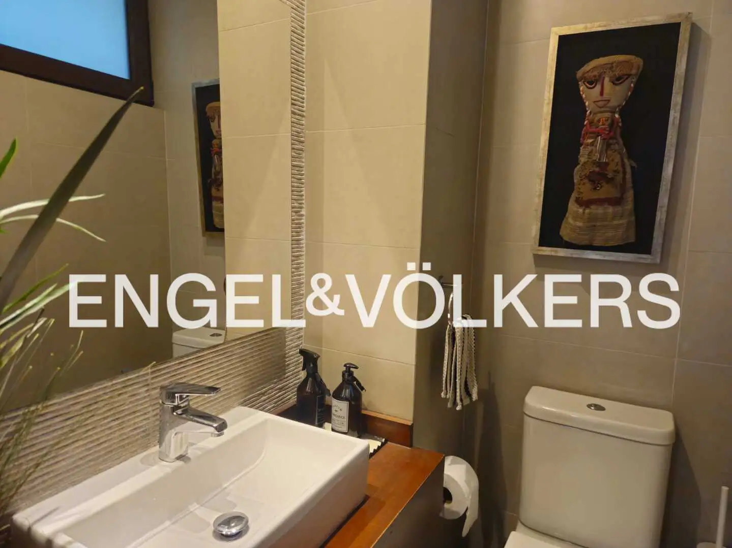 Modern bathroom with beige tile walls, a white sink and toilet, and a framed tribal art piece on the wall.