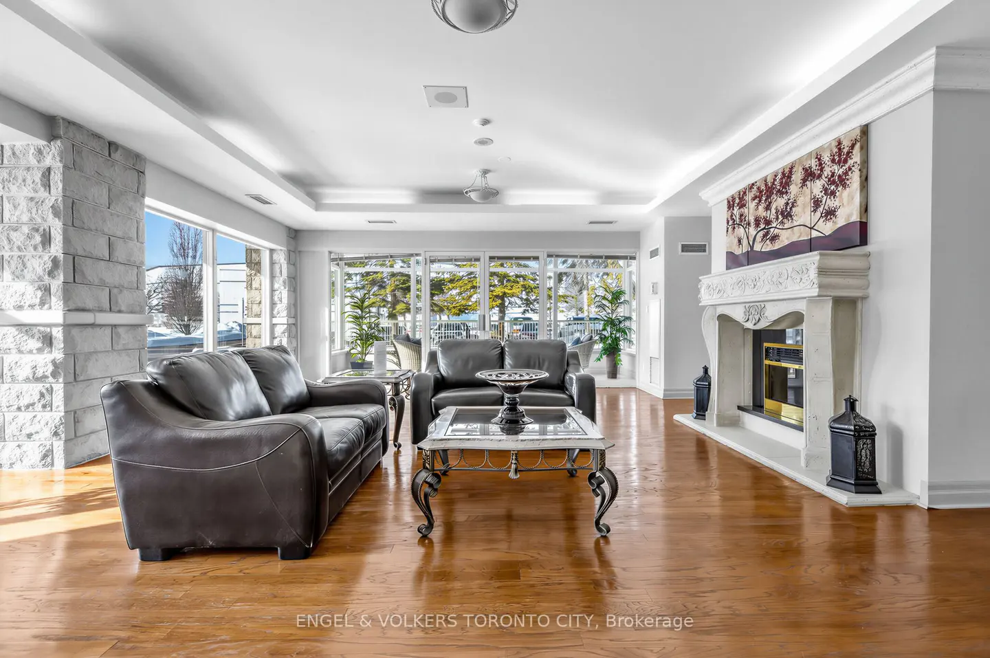 Bright living room with hardwood floors, two black leather sofas, a marble fireplace, and large windows.