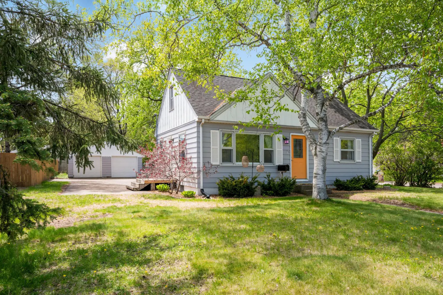 A gray, one-story house with a bright orange door and white trim, surrounded by green trees and grass. A detached garage is visible in the background.