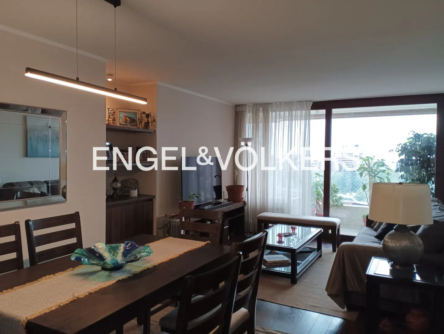 A wide shot of a living and dining area with dark wood furniture and a balcony with a view.