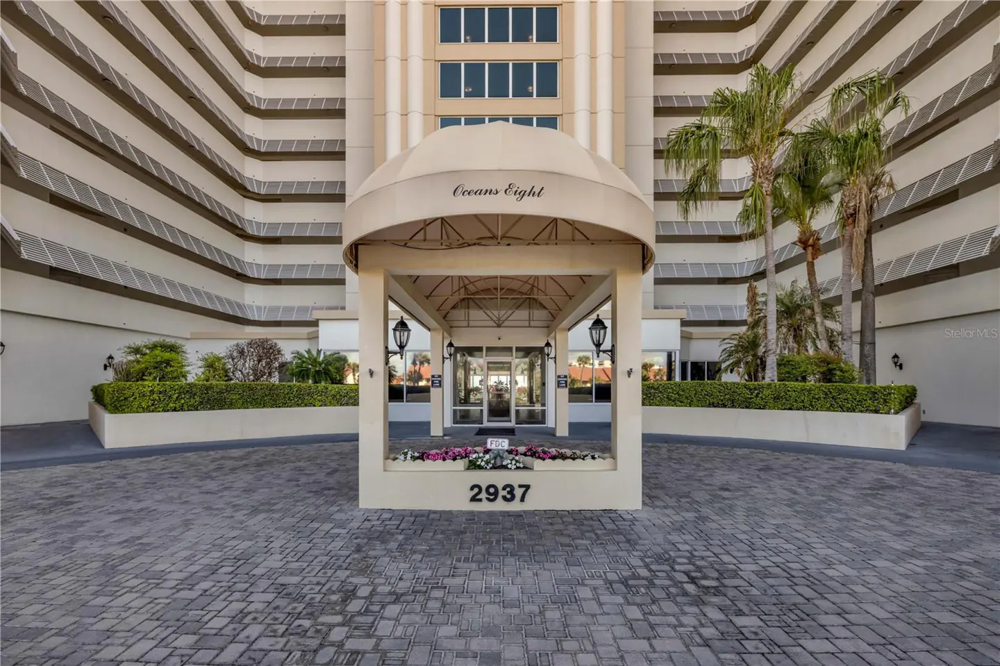 Exterior shot of Oceans Eight condo building entrance with beige awning, palm trees, and brick driveway.
