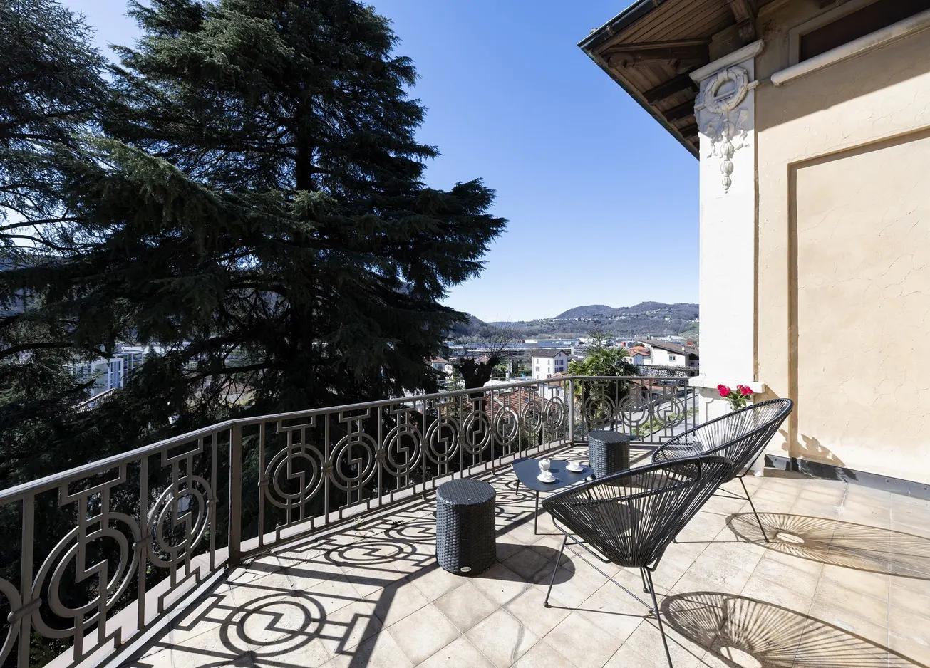 Balcony with black chairs and tables overlooks a city view. A large tree is visible to the left, and the sky is blue.