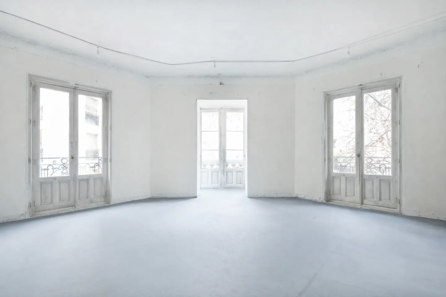 Bright, empty room with white walls and gray floor. Three sets of double doors with decorative iron balconies lead to outside.
