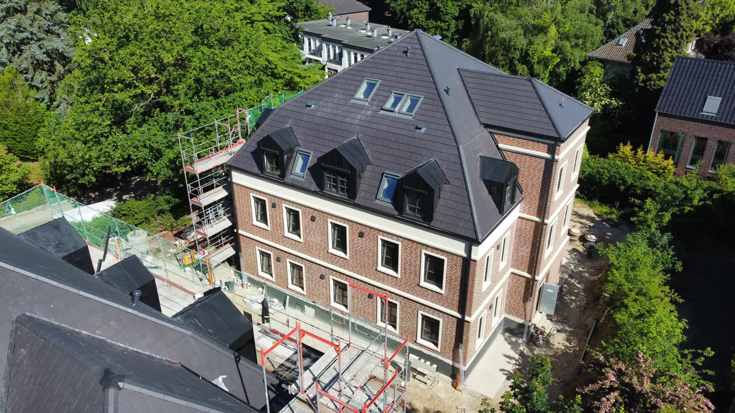 Aerial view of a three-story brick building with a dark gray roof and dormer windows, surrounded by green trees.