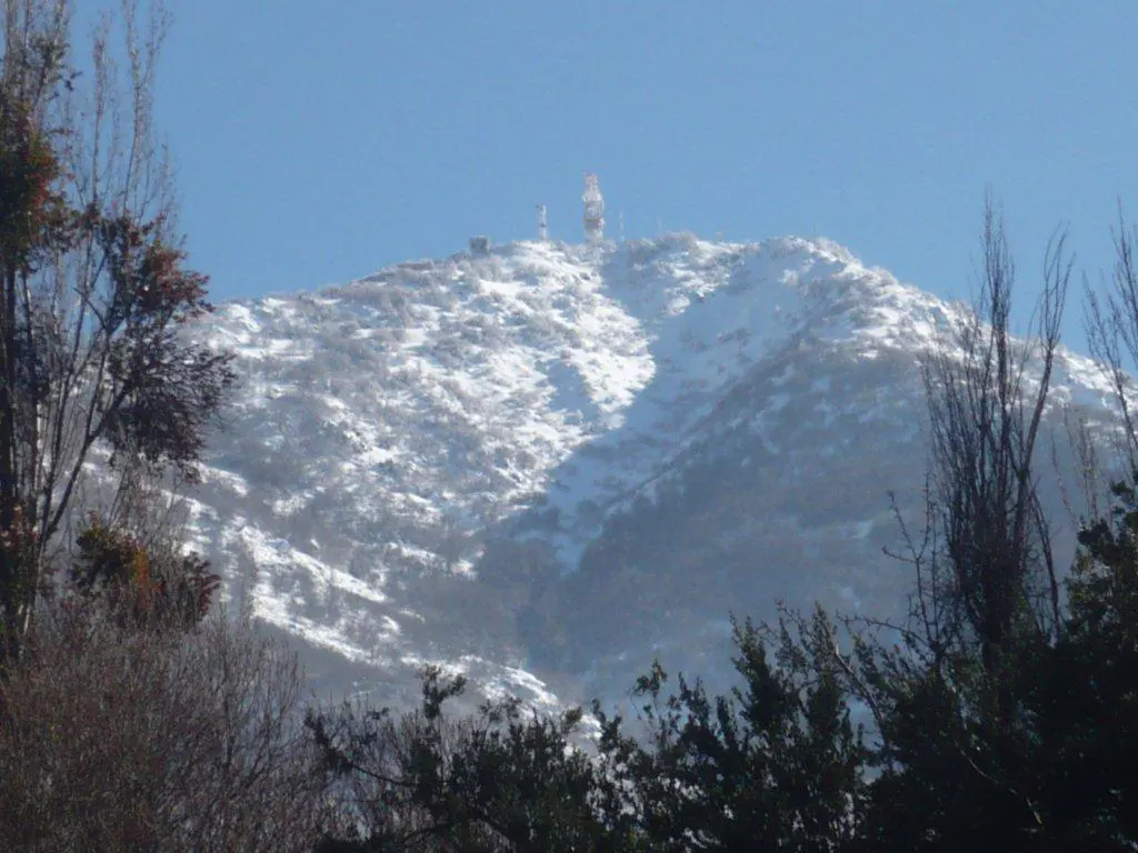 Snow-covered mountain with trees in the foreground and communication towers on top, under a clear blue sky.