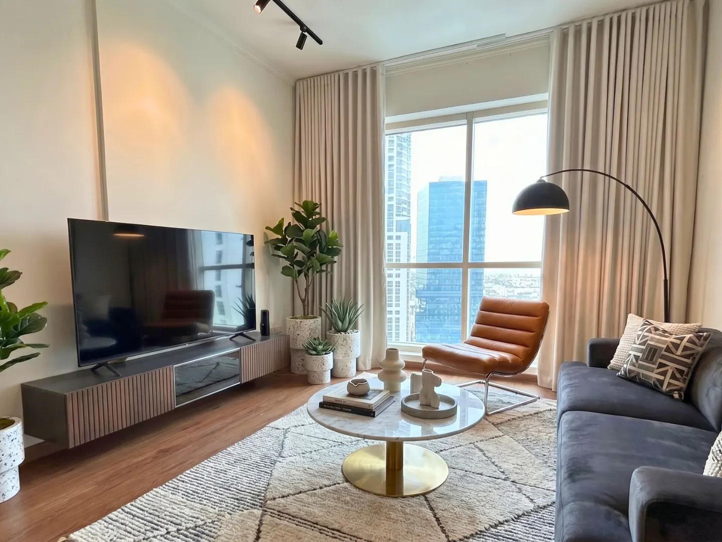 Living room with a TV, gray sofa, brown leather chair, and a large window with city views. Plants and a patterned rug add warmth.
