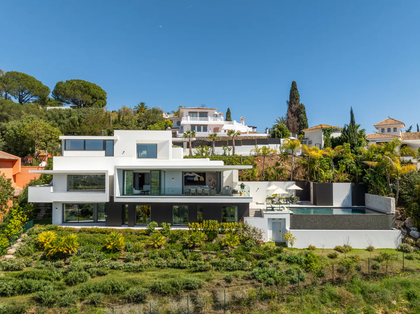 Modern white villa with a pool on a green hillside under a clear blue sky.