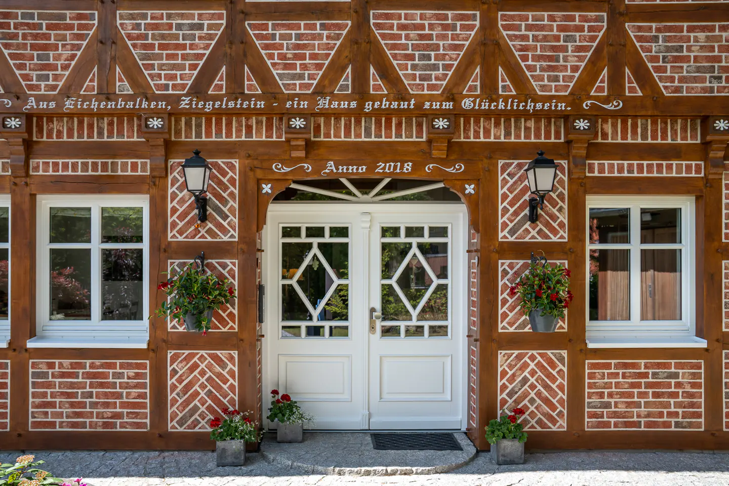White double doors with diamond-patterned glass on a half-timbered brick house with flower boxes and "Anno 2018" inscription.
