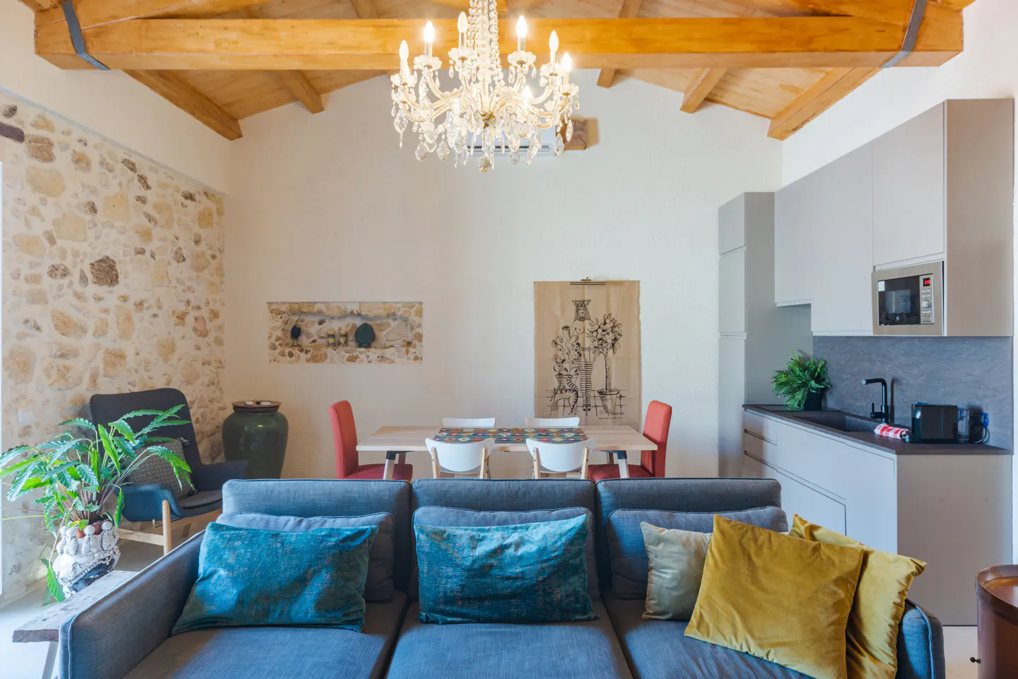 Living room with stone wall, wood beams, and chandelier. A gray sofa with colorful pillows sits in front of a dining table and kitchen area.