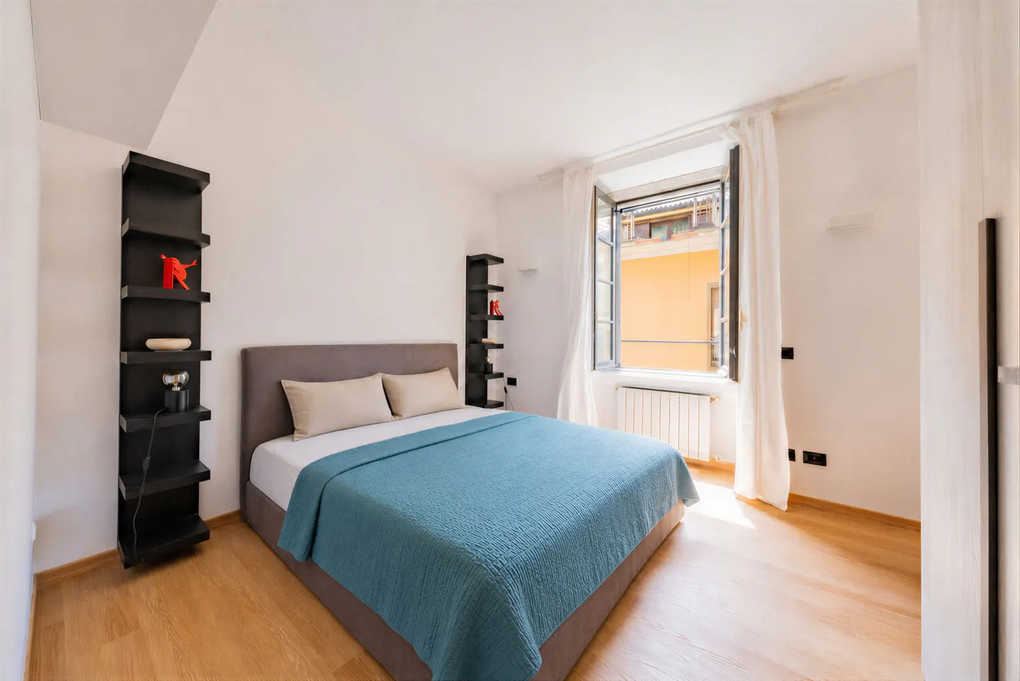 Bright bedroom with a grey bed, teal quilt, and two tall black shelves against white walls, featuring an open window.
