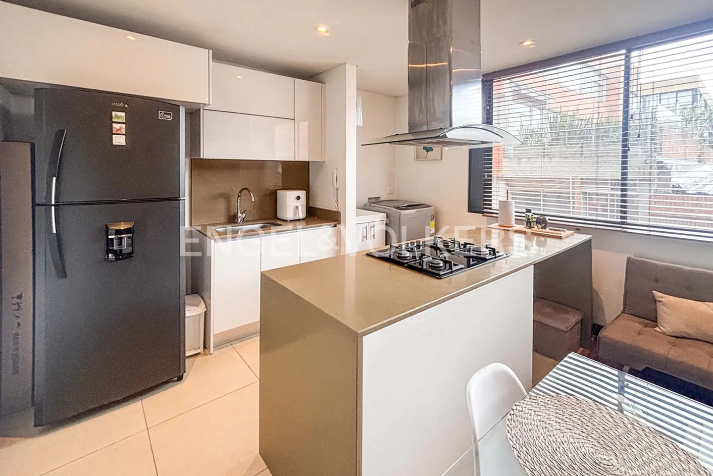 A modern kitchen with white cabinets, a stainless steel range hood, and a black refrigerator. A kitchen island with a cooktop and a dining area are visible.