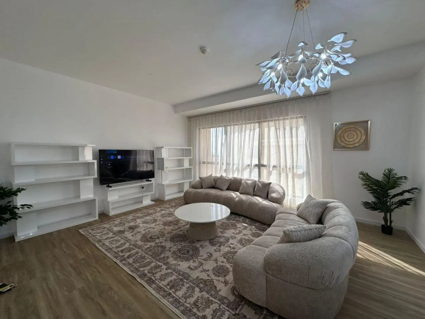 Bright living room with a curved beige sofa, white shelves, a TV, and a patterned rug. A modern chandelier hangs above a round white table.