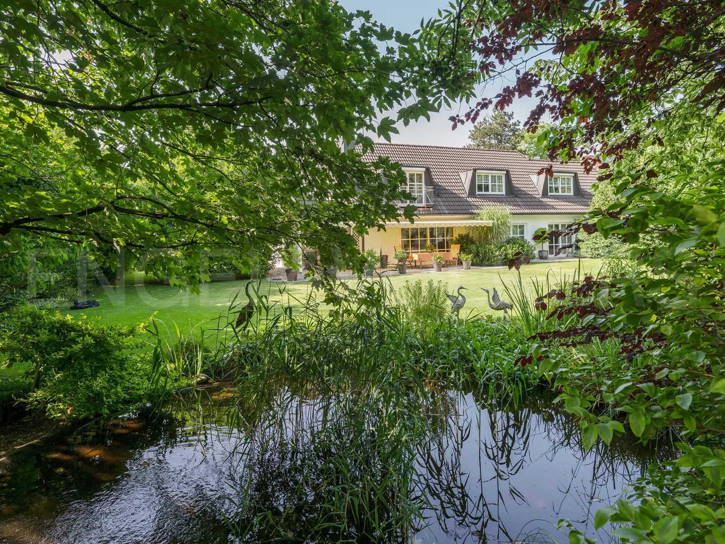 View of a white house with a brown roof, seen through green trees and a pond with heron statues.