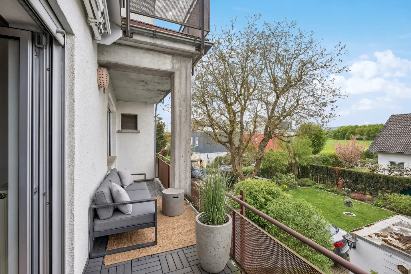 Balcony with gray sofa, table, and potted plant overlooking a green garden with trees under a blue sky.