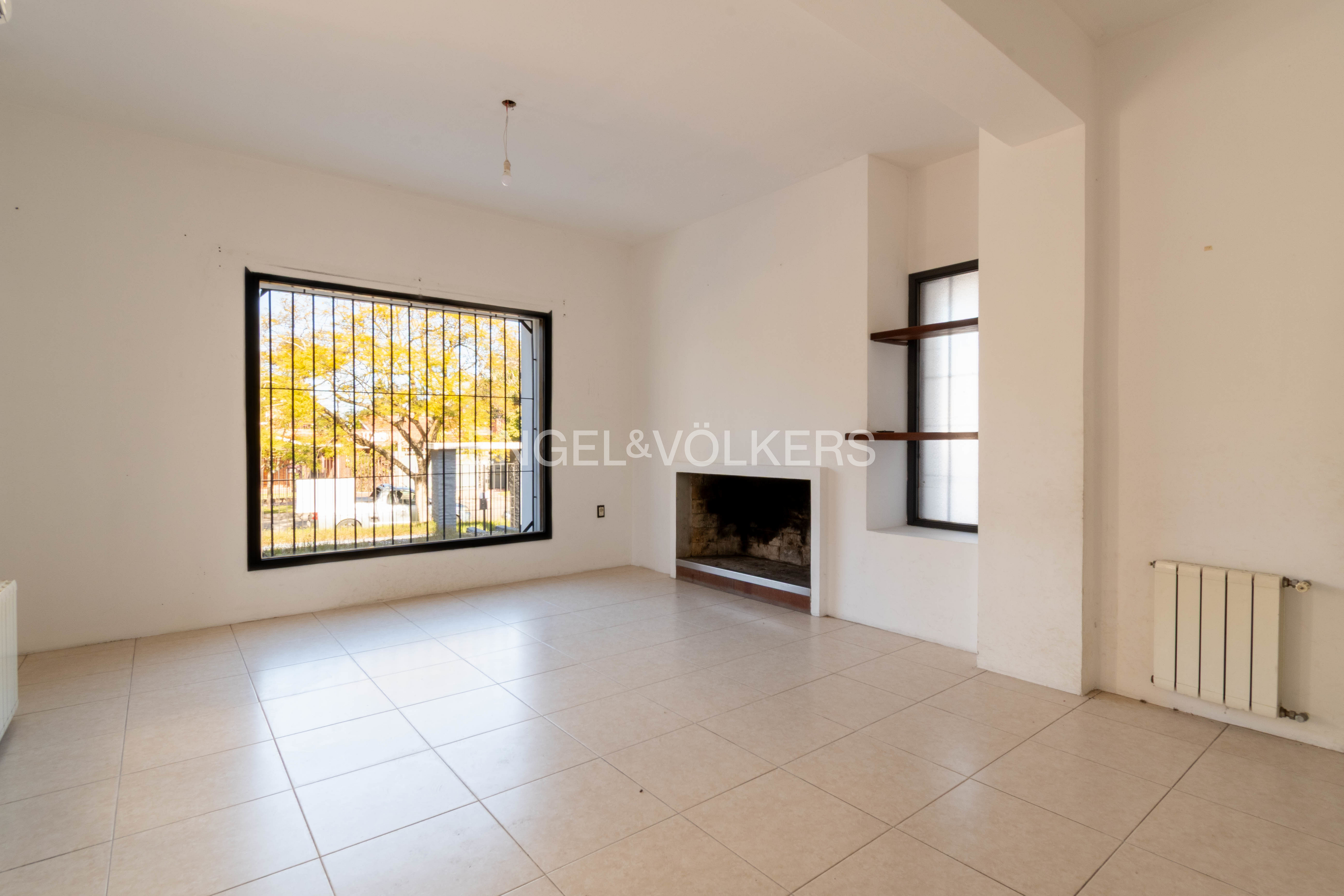 Empty living room with white walls, tiled floor, a fireplace, a large window with security bars, and a wall radiator.