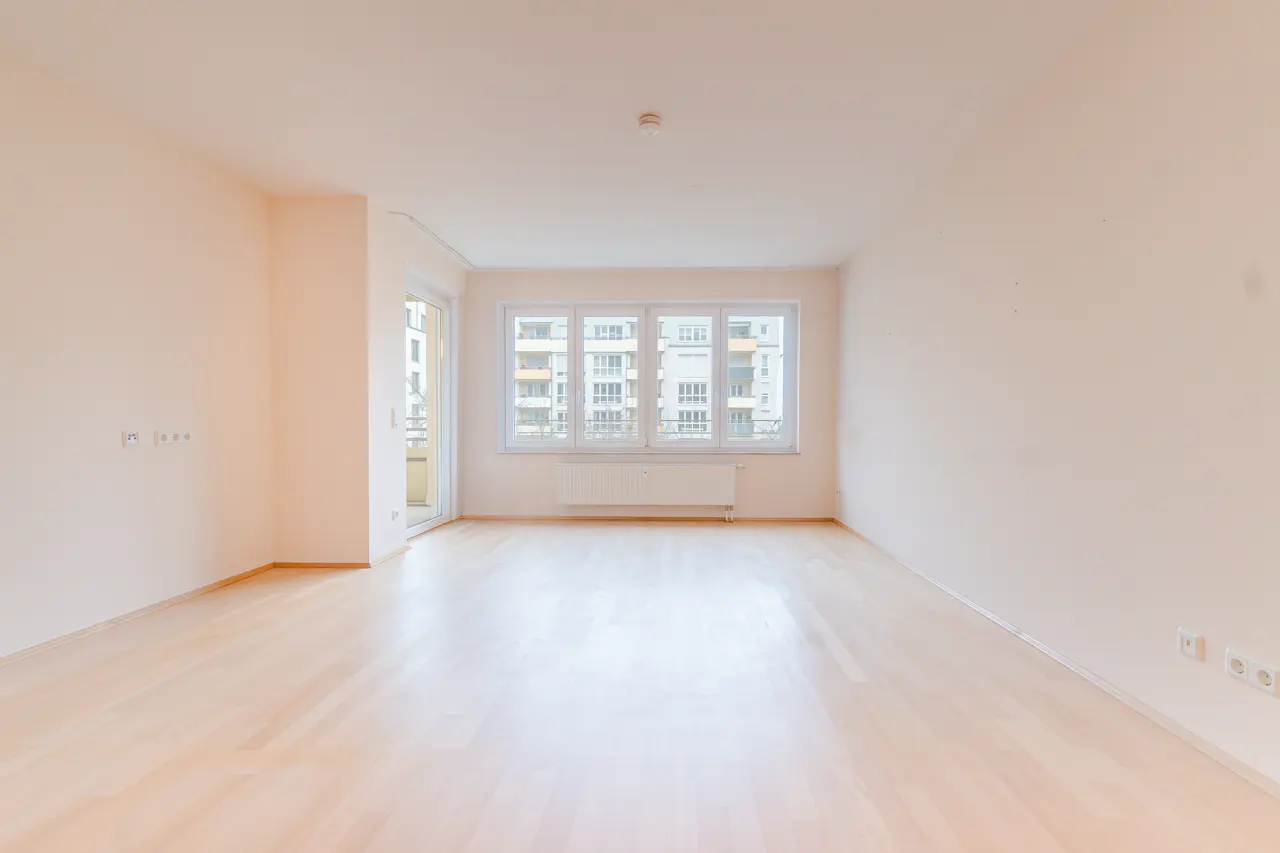 Bright, empty room with light wood floors and cream walls. Large window shows an apartment building. Balcony door on the left.