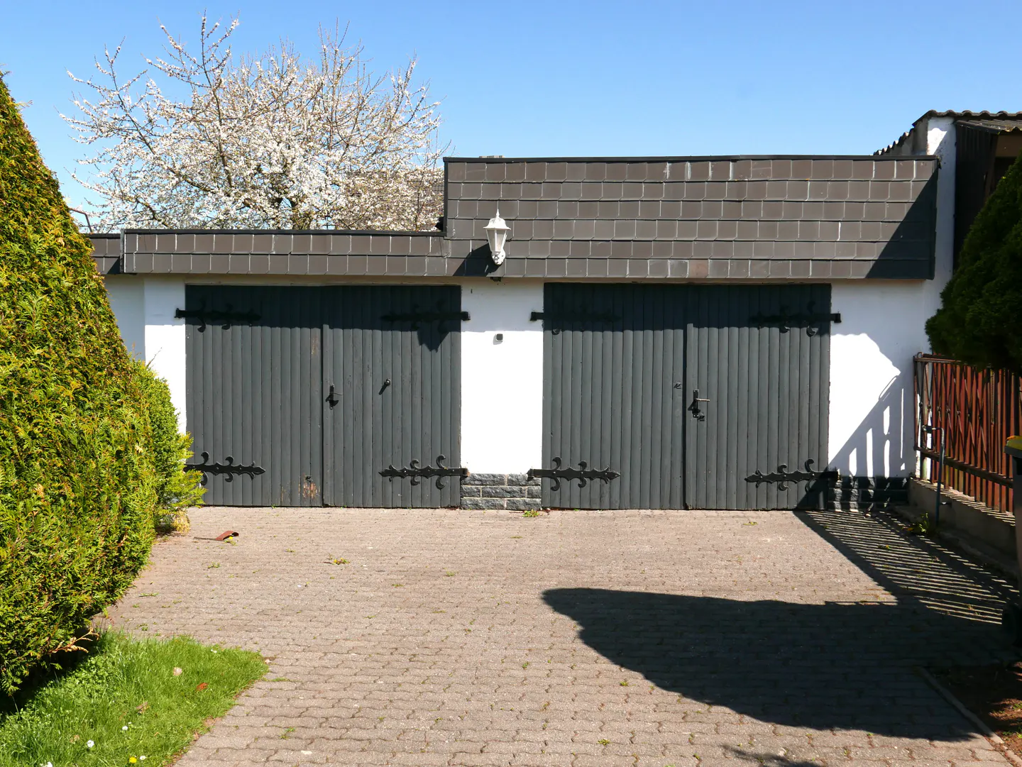 A two-car garage with gray doors and white walls, under a blue sky. A brick driveway leads to the garage.