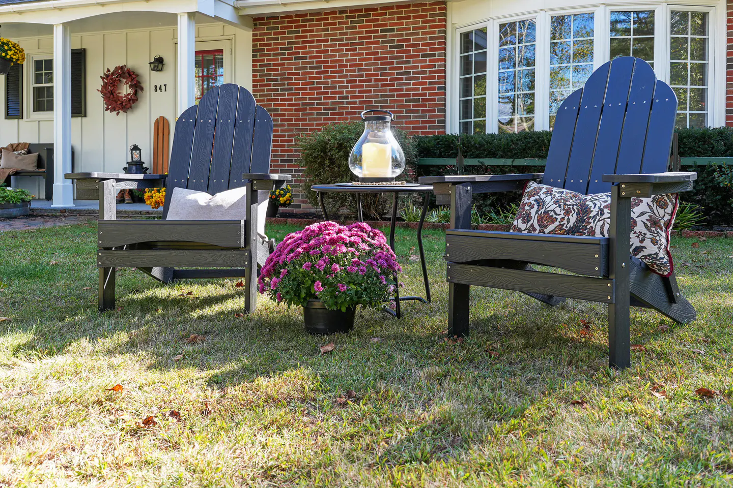 Two dark Adirondack chairs sit on a lawn with a small table, potted purple mums, and a house in the background.