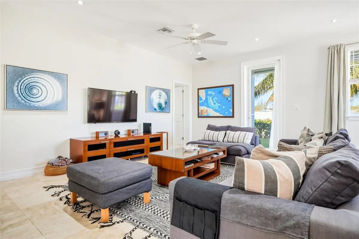 Bright living room with gray sofas, wood furniture, and coastal-themed art. A ceiling fan and large windows add to the airy feel.