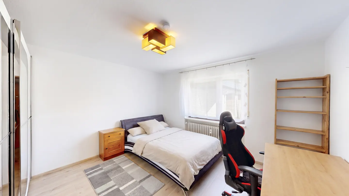 Bright bedroom with a bed, nightstand, desk, and gaming chair. A modern light fixture hangs from the ceiling.