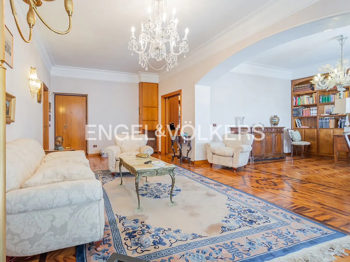 Elegant living room with white sofa, armchairs, and a blue patterned rug. A crystal chandelier hangs above a marble-topped coffee table.