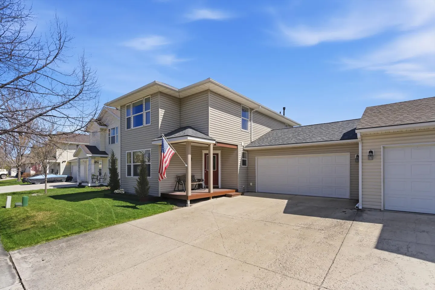 Two-story beige house with an American flag on the porch, a driveway, and a green lawn under a blue sky.